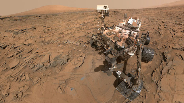 Photo autoportrait du rover Curiosity : l'astromobile est sur la droite avec en fond le sol martien et un mont au loin. Le rover possède des éléments blanc et noirs (notamment les roues) et est légèrement recouvert de poussière rouge martienne.