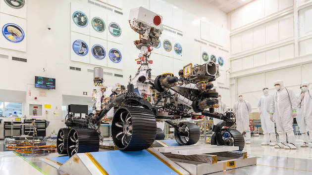 Le rover Perseverance en salle blanche pendant une phase de test : on voit ses six roues sur deux tapis bleu qui simulent le sol accidenté de Mars et sur sa droite se tiennent cinq ingénieurs en combinaison.  