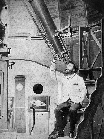 Photo en noir et blanc de Camille Flammarion, assis en blouse blanche en train d'observer dans sa lunette astronomique, pointée au-dessus de lui vers le ciel, au sein de la coupole de son observatoire. 