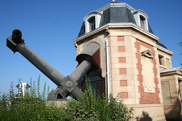 Bâtiment historique de l'Observatoire de Lyon, en briques rose clair et blanches, toit noir, d'où dépasse la lunette coudée de couleur gris anthracite. En fond le ciel est bleu. 