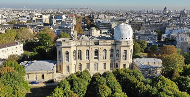 Vue en survol du bâtiment de l'Observatoire de Paris, en pierres blanches avec de grandes fenêtres. Il est entouré de verdure, et en fond on voit les toits de Paris. 