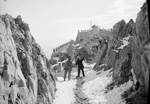 Photo en noir et blanc qui montre le site du Pic du Midi au début du XXe siècle. On voit à droite et à gauche une paroi rocheuse, au centre un chemin enneigé avec deux hommes debout regardant la roche, et derrière les premiers bâtiments de l'Observatoire. 