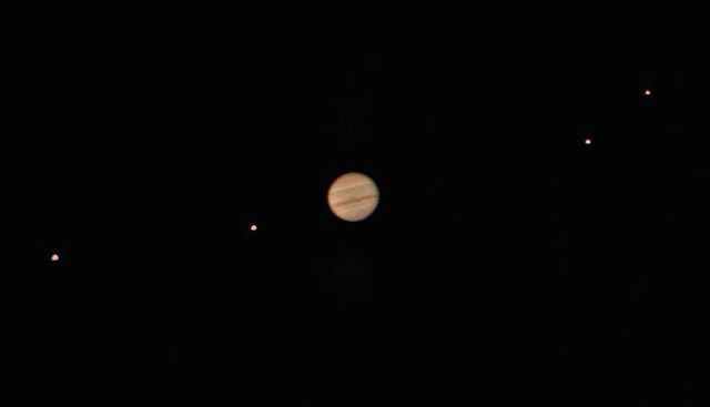 Sur fond noir, Jupiter au centre dont on distingue l'atmosphère aux motifs horizontaux en couleur marron clair, et quatre petits points, deux de chaque côté de la planète qui l'entourent. Ce sont ses quatre lunes. 