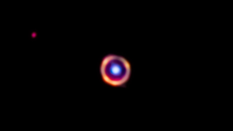 Sur un fond noir, il y a deux objets remarquables : une galaxie au premier plan vue comme un gros point bleu vif au centre, qui est entouré d'un anneau orange montrant la découverte de molécules organiques. Près du coin supérieur gauche de l'image, il y a aussi une galaxie d'arrière-plan lointaine représentée par un petit point rouge.