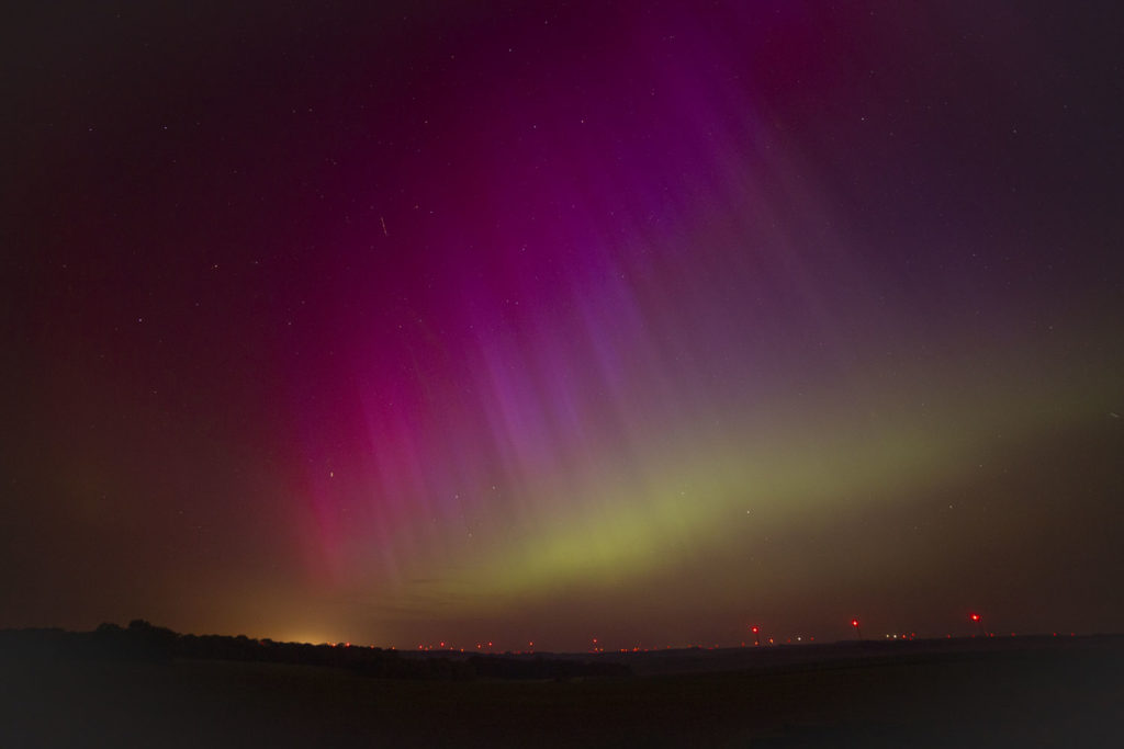 Photo grand angle prise sur un espace dégagé la nuit, où l'on voit des lumières rouges d'éoliennes à l'horizon. Dans le ciel, l'aurore boréale couvre tout le ciel, elle est verte près de l'horizon et a un beau dégradé de mauve et de rouge plus haut dans le ciel. Il y a de nombreux piliers lumineux.