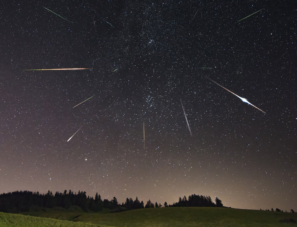 Image montrant des étoiles filantes qui semblent toutes provenir d'un même point dans le ciel étoilé, situé dans la constellation de Persée. On voit un ciel étoilé, de nombreux traits blancs qui correspondent aux étoiles filantes et un premier plan de collines et de sapins.