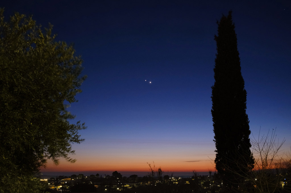 Photo d'un ciel crépusculaire bleu foncé, avec un liseré de rouge et d'orange le long de l'horizon. En premier plan, un arbre feuillu à gauche et un sapin à droite. A l'horizon, un village éclairé.