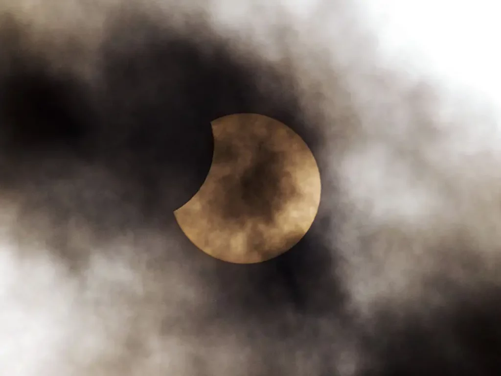 Photo montrant une éclipse le Soleil avec une échancrure sombre en haut à gauche (la Lune passe devant) et partiellement voilé par des nuages légers.